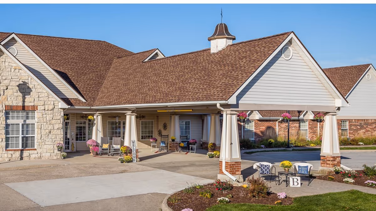 Front entrance of a single-story senior living building with a covered porte-cochère, columns, seating, and flower planters.