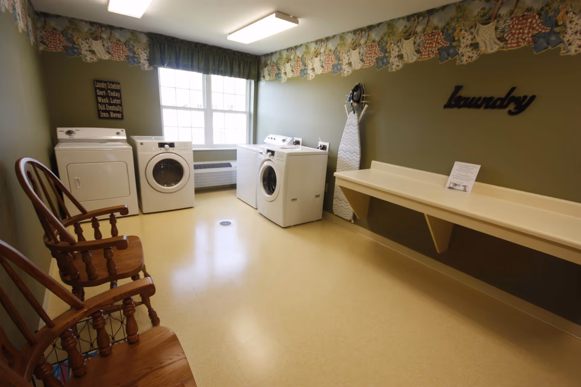 A clean laundry room with multiple washers and dryers, a long folding counter, wooden chairs, and an ironing board beneath a window.