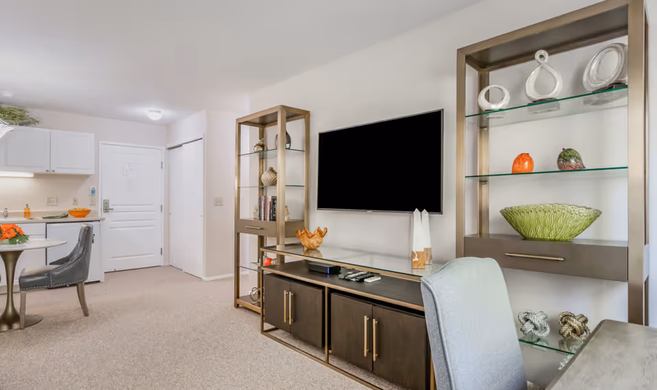 Interior view of a senior living community apartment featuring a small dining area with a round table and two chairs, a kitchenette with white cabinets and a countertop, a wall-mounted flat-screen TV above a dark wood media console, and decorative shelving units with various ornaments and glass shelves.