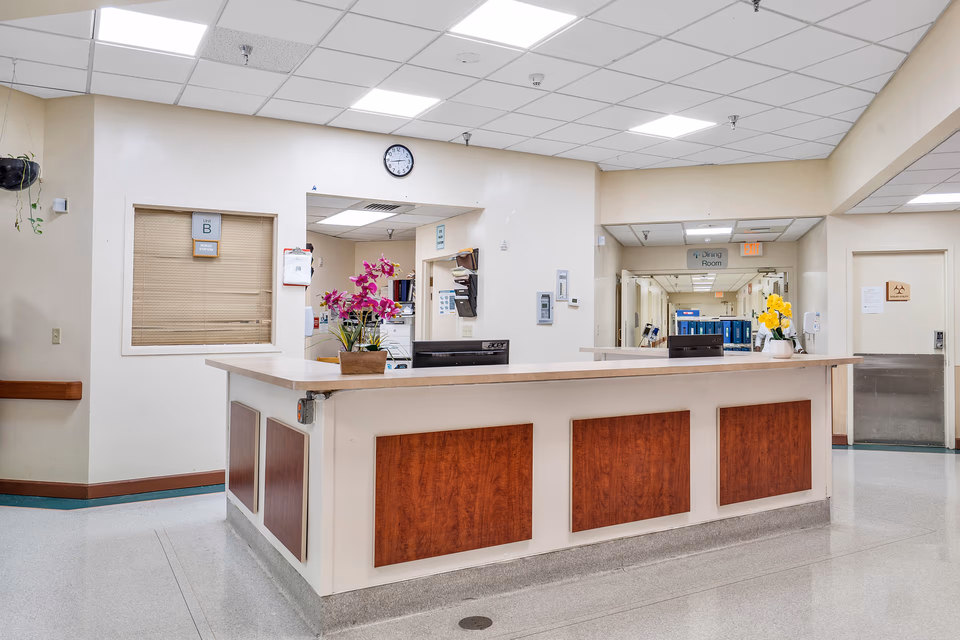 Reception desk area inside Shoreside Health and Rehabilitation Center with a wooden counter, two flower pots with purple and yellow flowers, a clock on the wall, and a hallway leading to other rooms including a dining room.