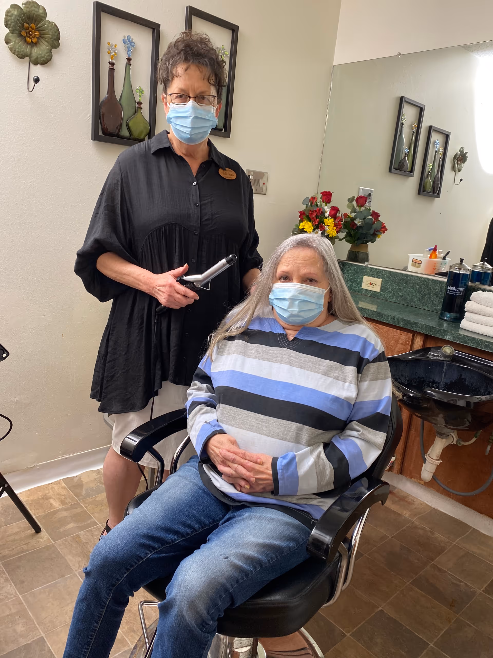 A woman wearing a striped sweater and a face mask is seated in a salon chair while another woman, also wearing a face mask and holding a hair styling tool, stands behind her in a salon setting with a sink, mirror, and floral decorations in the background.