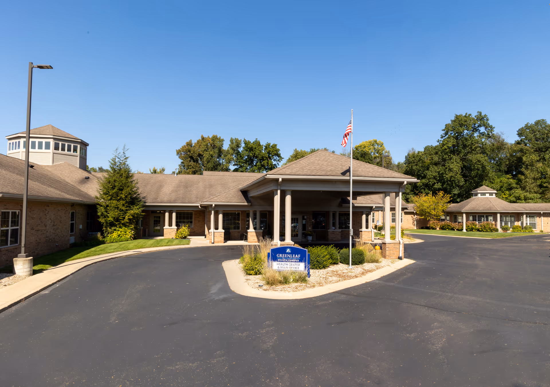 Front entrance of Greenleaf Health Campus with a covered porte-cochère, flagpole, sign, and a landscaped circular driveway under a clear blue sky.