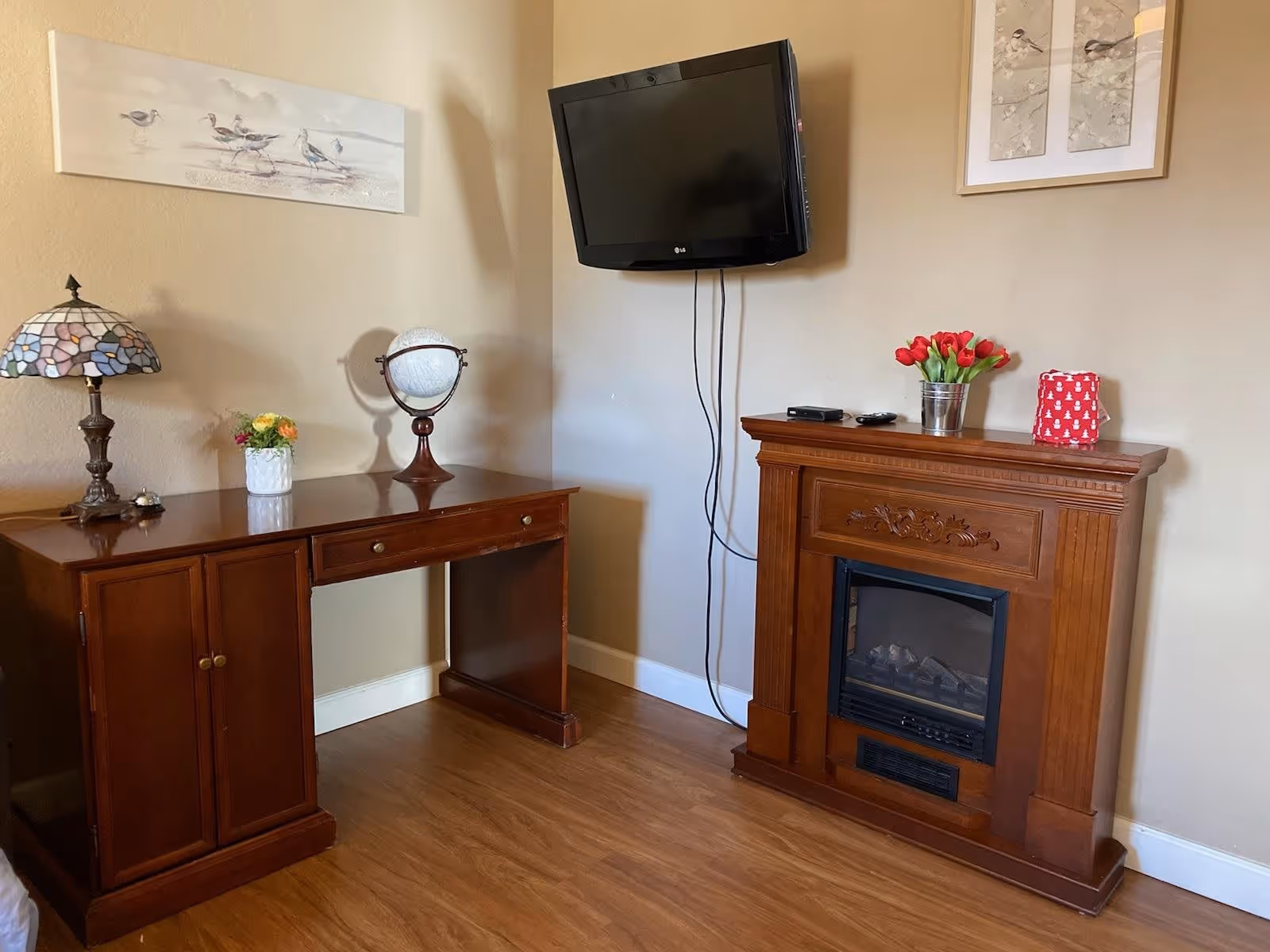 A cozy corner of a room featuring a wooden desk with a Tiffany-style lamp, a small flower vase, and a globe. Mounted on the wall above is a flat-screen TV. Next to the desk is a wooden electric fireplace mantel decorated with a small bucket of red tulips and a red gift bag. The walls are beige with two framed artworks hanging.