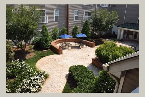 Outdoor courtyard area with paved walkways, green bushes, trees, and a seating area featuring tables with blue umbrellas surrounded by a low brick wall. The courtyard is adjacent to a multi-story building with several windows.