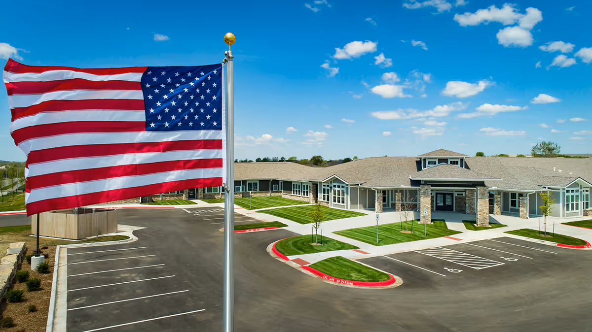 An American flag flies in front of a single-story assisted living facility with its parking lot and entrance under a blue sky.