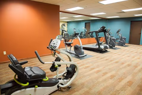 Exercise room with recumbent bike, treadmills and ellipticals against a mirrored wall and orange accent wall.