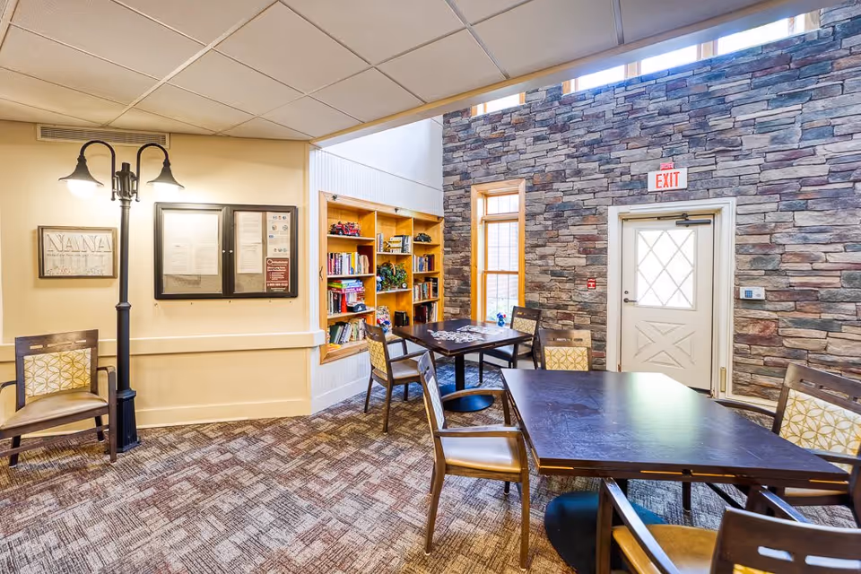 A cozy common area in a senior living facility with a stone accent wall, a door with a window, and a large window letting in natural light. The room features a dark wooden table with several chairs around it, a smaller table with chairs near a built-in bookshelf filled with books and decorative items. A floor lamp with two lights and a framed sign that says 'NANA' are visible on the left wall. The carpet has a patterned design, and the ceiling has a grid pattern with recessed lighting.