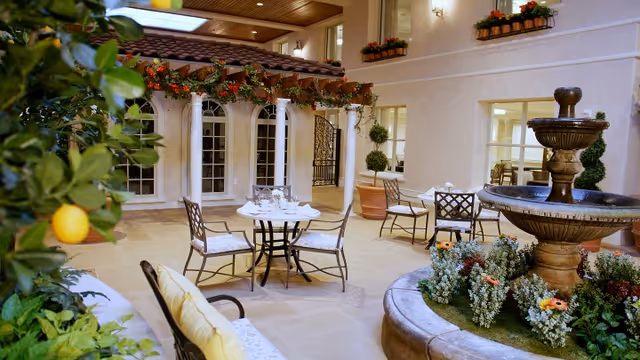 Indoor courtyard area with a round table and chairs, a water fountain surrounded by flowers, potted plants, and a pergola decorated with hanging flower baskets. The space has a warm, inviting atmosphere with large windows and a wooden ceiling.