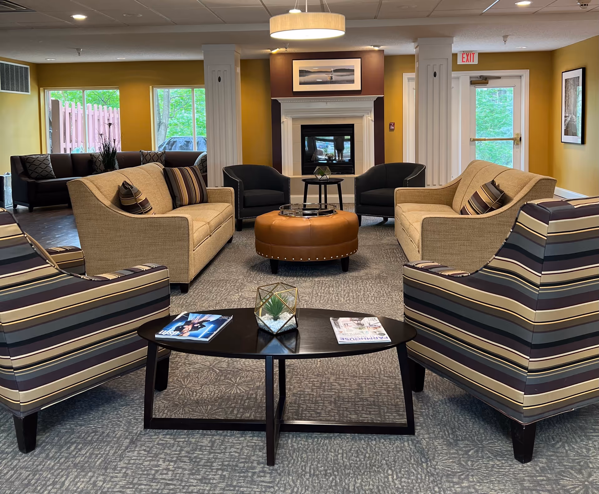 A cozy living room area with two beige sofas, two striped armchairs, and two black armchairs arranged around a round brown ottoman and a small black coffee table with magazines and a decorative plant. The room has yellow walls, large windows showing greenery outside, a fireplace with a framed picture above it, and an exit door.