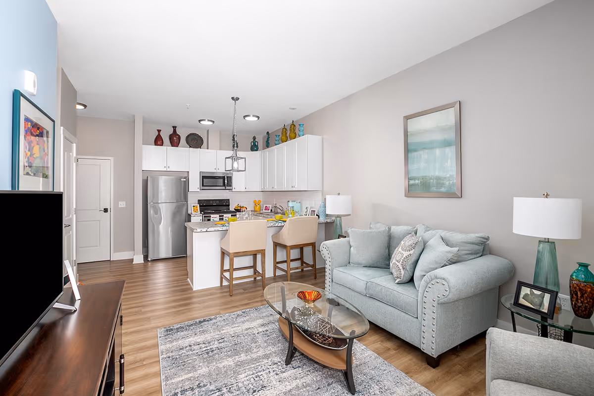 Open-concept living room and kitchen with a light-blue sofa, glass coffee table, island with bar stools, and stainless steel appliances.