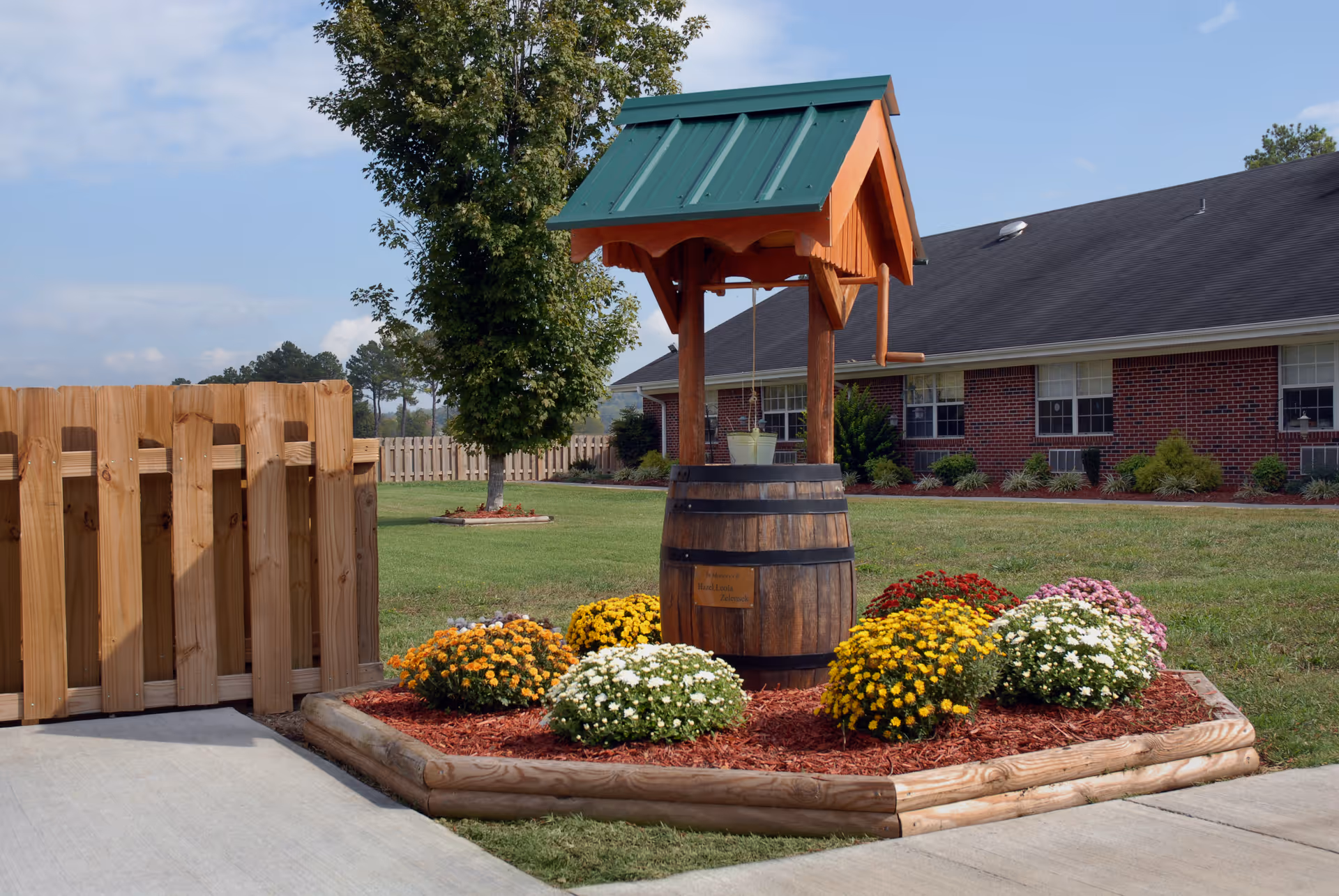 A decorative wishing well surrounded by colorful flowers in a landscaped garden area with a wooden fence and a brick building in the background under a partly cloudy sky.