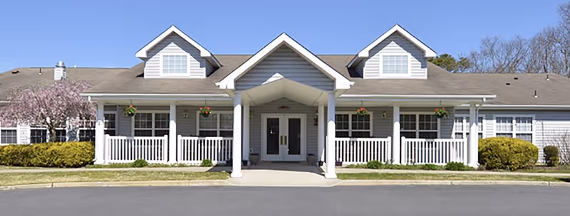 Front exterior view of a single-story senior living facility building with a covered entrance supported by white columns, white railings, and a well-maintained lawn with bushes and a flowering tree under a clear blue sky.