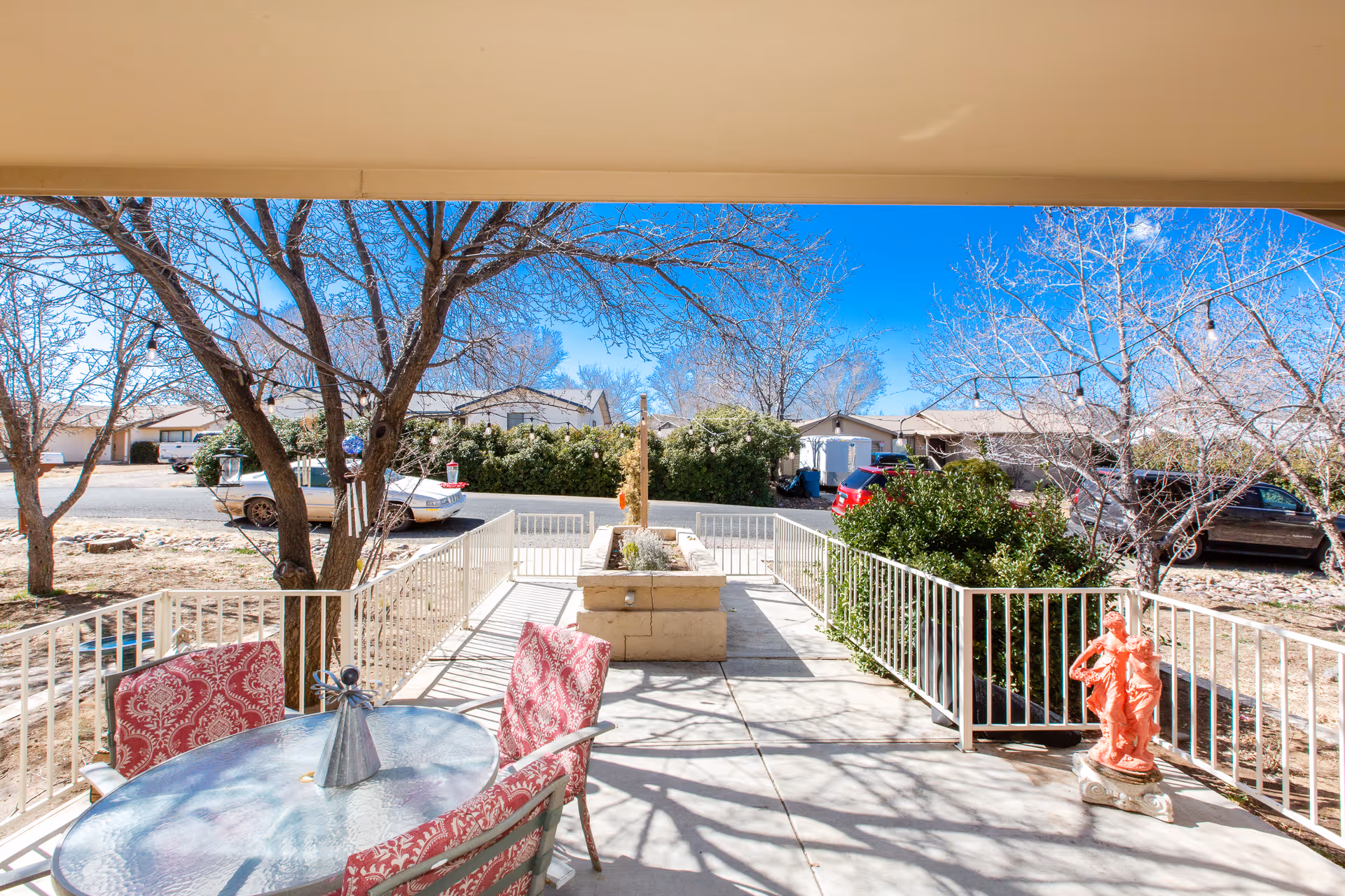 Outdoor patio area with a glass table and four red patterned chairs. The patio is surrounded by white metal railings and has a planter box in the center. There are leafless trees and bushes around the patio, with a clear blue sky and residential houses in the background.