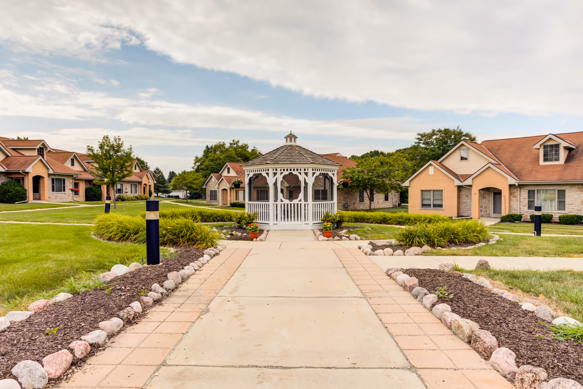 A paved walkway leads to a white gazebo in the center of a landscaped courtyard with green grass, shrubs, and trees. Surrounding the courtyard are single-story residential buildings with beige walls and reddish-brown roofs under a partly cloudy sky.
