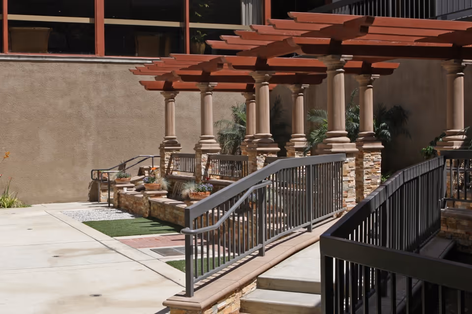 Outdoor patio area with a pergola supported by stone columns, benches, potted plants, and a ramp with metal railings leading up to the seating area.