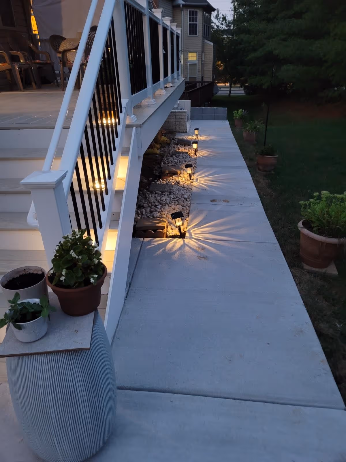 Outdoor walkway beside a building with a white railing and stairs. The walkway is lined with small garden lights casting star-shaped patterns on the concrete path. Several potted plants are placed along the walkway and near the stairs. It is dusk or early evening with some lights on inside the building.
