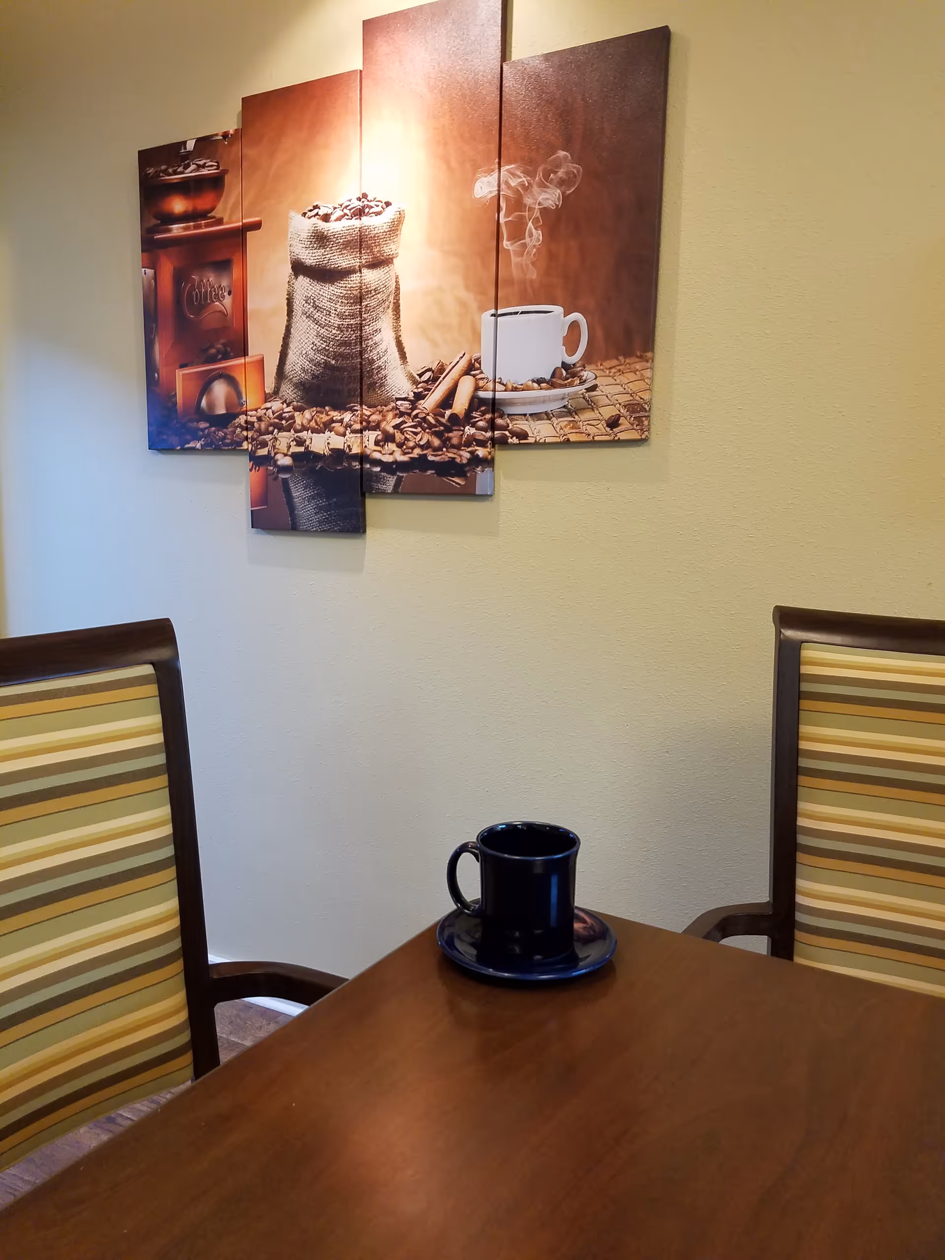 A wooden table with a dark blue coffee cup and saucer on it, flanked by two striped cushioned chairs. On the wall behind the table is a multi-panel artwork depicting a burlap sack filled with coffee beans, cinnamon sticks, and a steaming cup of coffee.