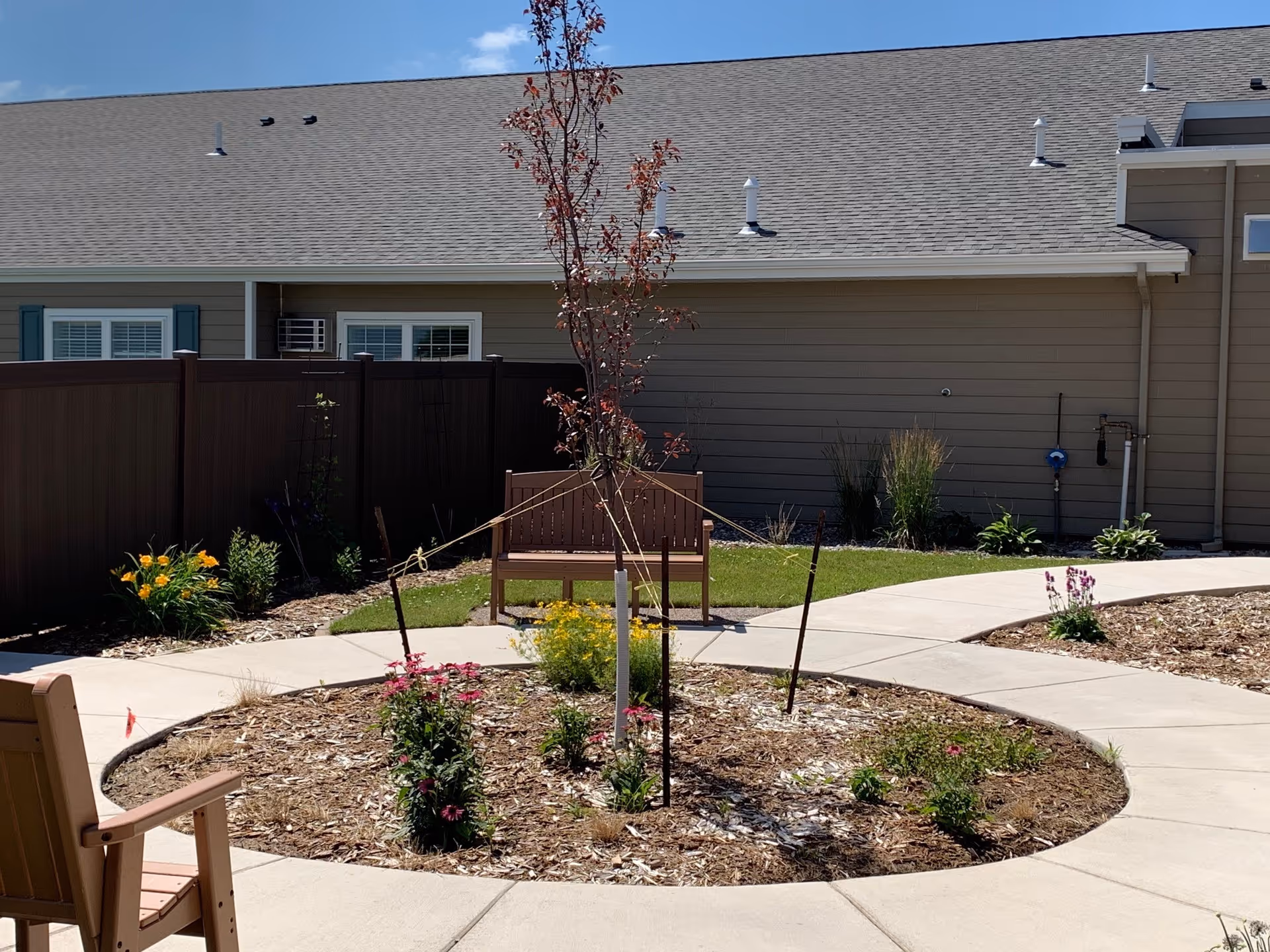 Outdoor garden area with a circular concrete walkway surrounding a mulched flower bed with small plants and a young tree supported by stakes. There are wooden benches placed along the walkway, a brown fence, and a beige building with windows and a gray roof in the background under a clear blue sky.