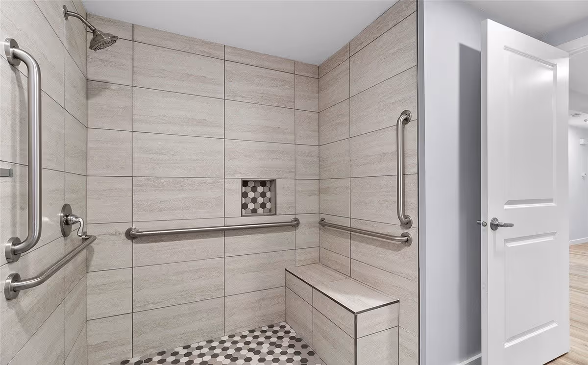 A modern accessible shower area with beige tiled walls and hexagonal patterned floor tiles. The shower has multiple stainless steel grab bars and a built-in bench. A white door is open leading to a hallway with light wood flooring.