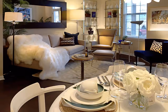 A cozy living room area with a beige sofa draped with a white fluffy throw, a beige armchair with an ottoman, a dark blue chair, and a round wooden coffee table. The room is warmly lit with floor lamps and natural light from windows. In the foreground, a dining table is set with plates, bowls, glasses, and a centerpiece of white flowers. Shelves with decorative plants and books are visible in the background.