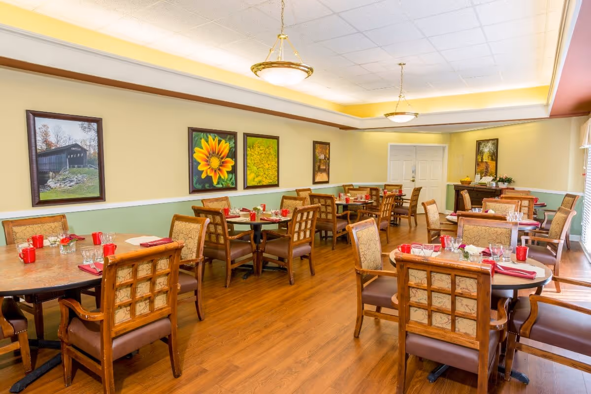 A dining room with multiple round and rectangular tables set with red napkins, glasses, and small flower arrangements. The room has wooden flooring, beige and green walls, framed pictures including a sunflower and a covered bridge, and ceiling lights with gold accents.