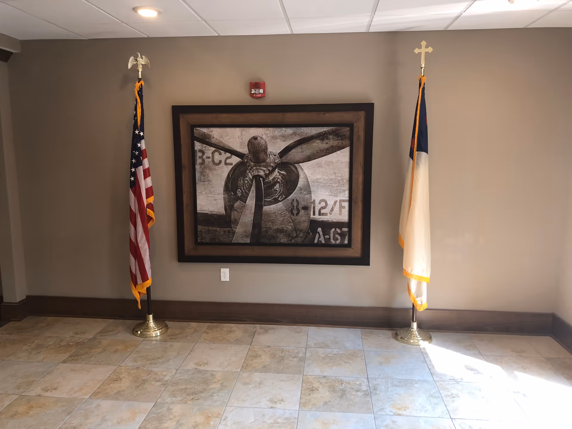 An interior wall with two flags on stands placed on either side of a framed black and white picture of an airplane propeller. The floor is tiled with beige and light brown tiles, and the walls are painted a light brown color.