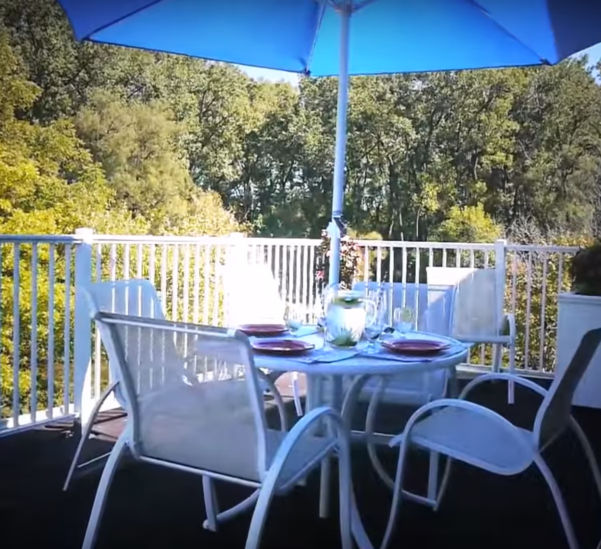 Outdoor patio area with a round glass table set for four, white chairs, a blue umbrella providing shade, and a background of green trees.