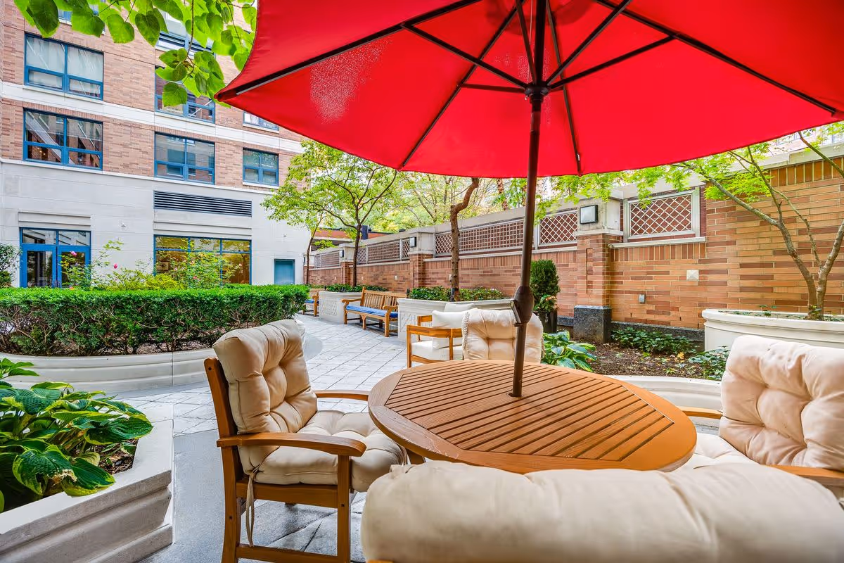 Outdoor patio area with cushioned wooden chairs around a wooden table with a large red umbrella. The space is surrounded by greenery, trees, and brick walls with a building visible in the background.