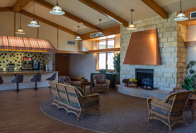 A cozy common area with wicker chairs and a sofa arranged around a stone fireplace with a copper hood. The room has wooden beams on the ceiling, hanging pendant lights, and a small counter area with bar stools and a coffee machine under a striped awning. Large windows let in natural light, and there are plants and small tables with flowers adding to the welcoming atmosphere.