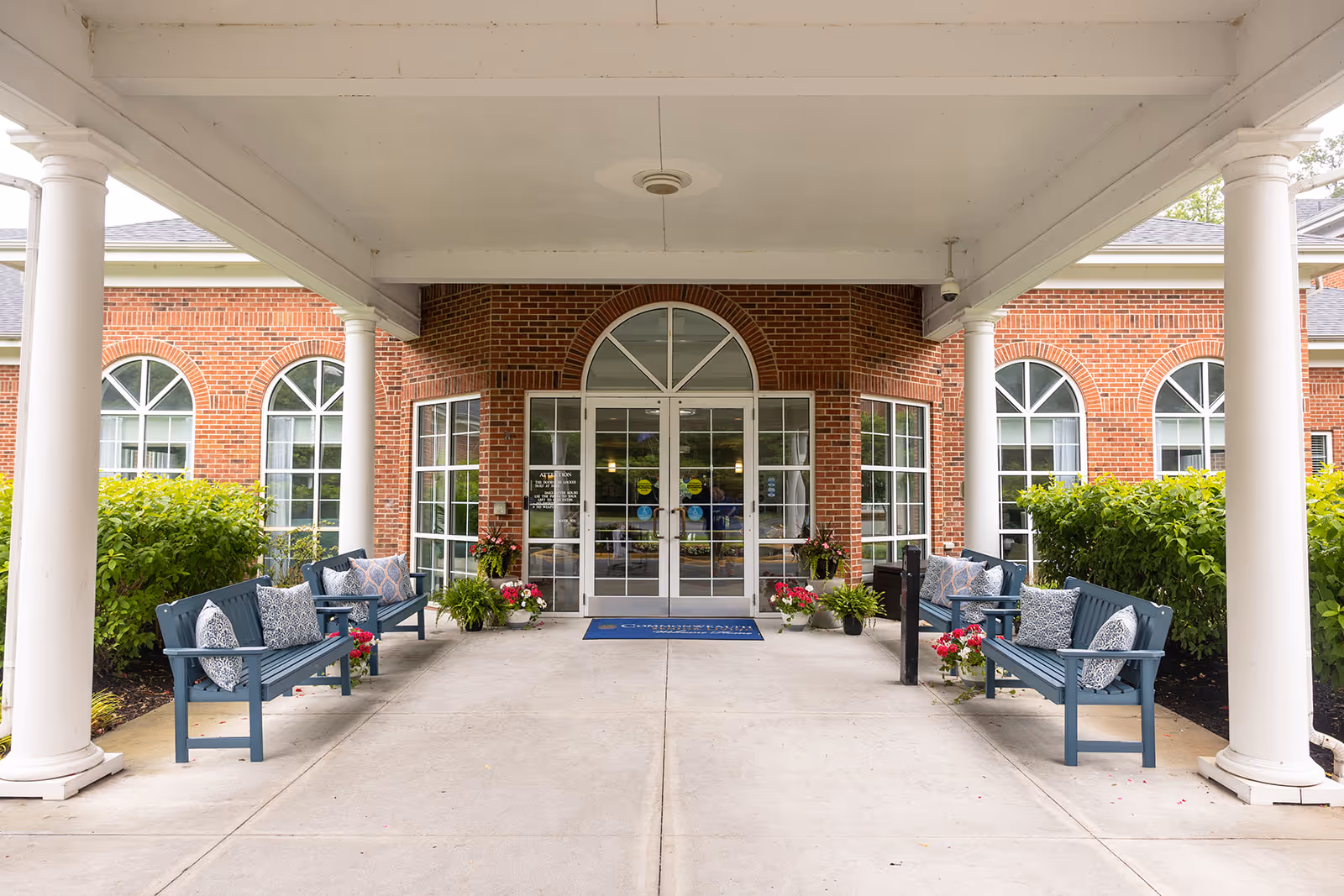 Covered entrance area of Commonwealth Senior Living at Cedar Manor with four blue benches adorned with patterned cushions, potted plants with flowers, white columns supporting the roof, and glass double doors leading into a brick building with arched windows.