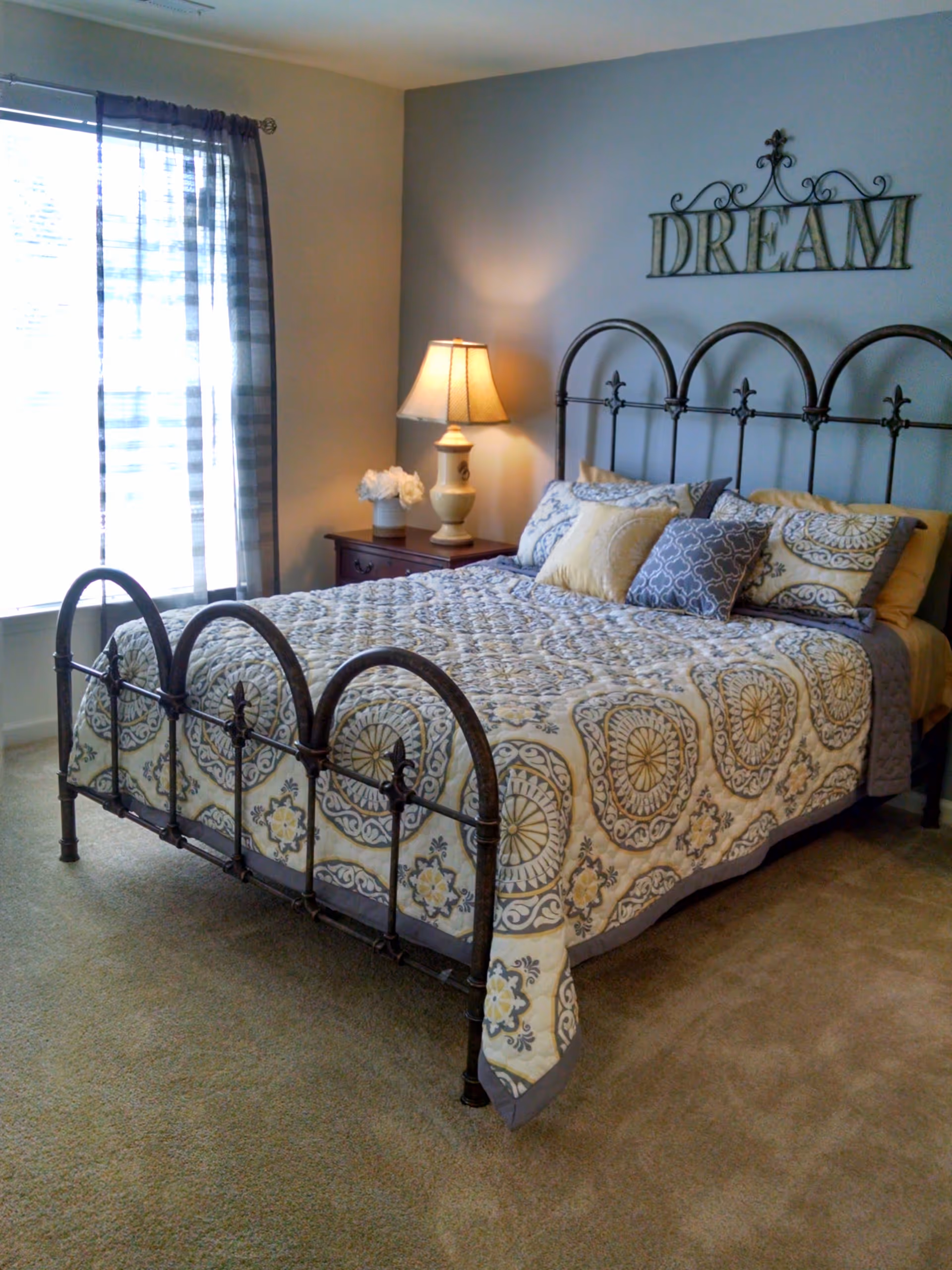 A cozy bedroom with a metal framed bed adorned with a patterned quilt and multiple pillows. A wooden nightstand beside the bed holds a lamp and a vase with white flowers. Light filters in through a window with sheer curtains, and a decorative metal wall art piece spelling 'DREAM' hangs above the bed.