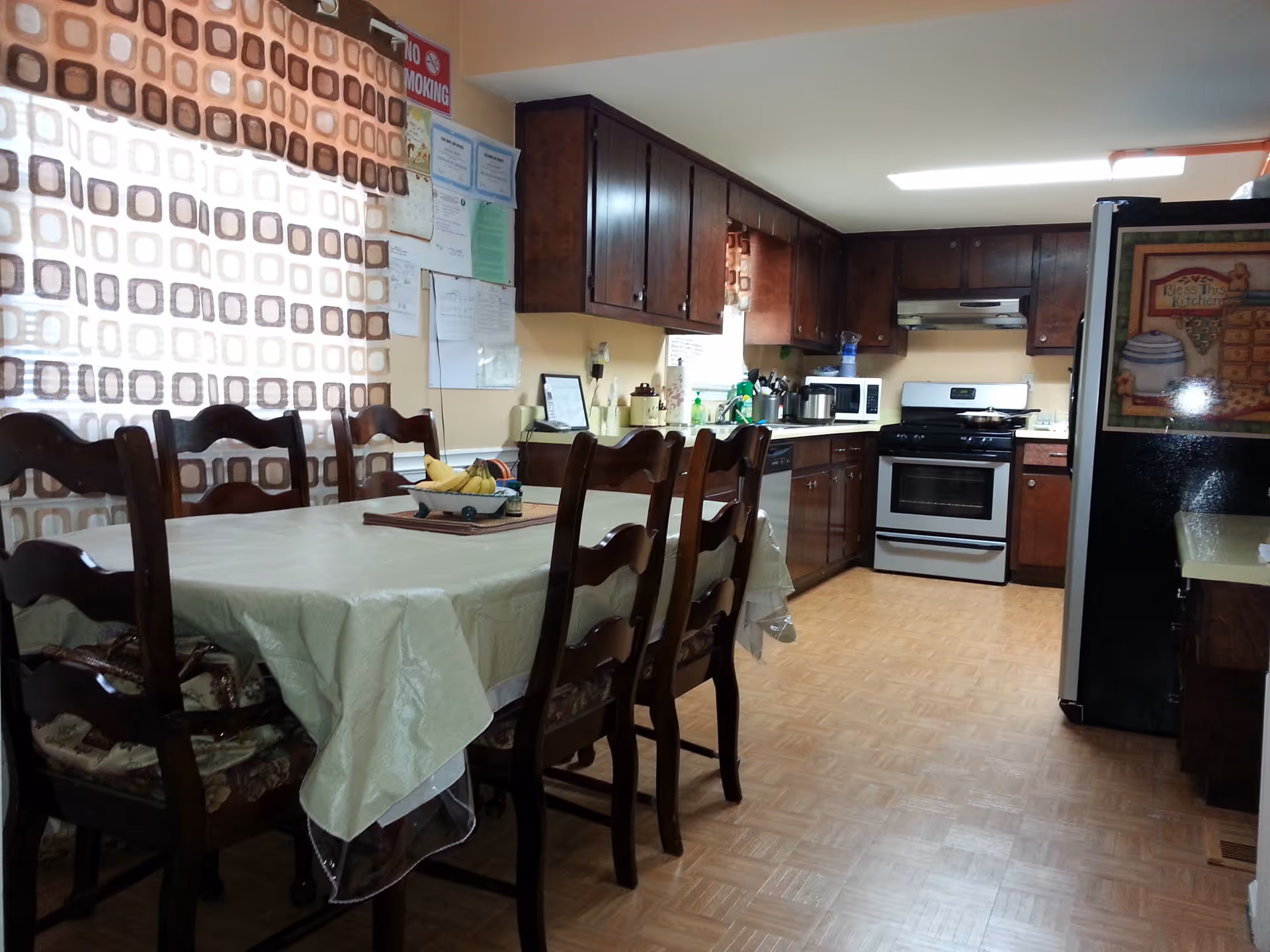 A kitchen and dining area with a table covered by a plastic tablecloth and six wooden chairs. The kitchen has dark wooden cabinets, a stove, microwave, and refrigerator. A window with patterned curtains is visible, along with various kitchen items on the counters.