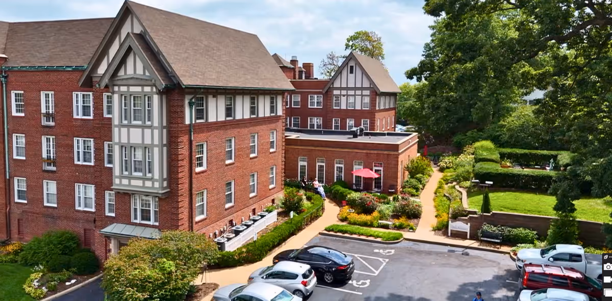 Exterior view of The Rockwood senior living facility showing a large brick building with multiple windows and a gabled roof. There is a parking lot with several cars in front, landscaped garden areas with pathways, benches, and greenery surrounding the building under a partly cloudy sky.