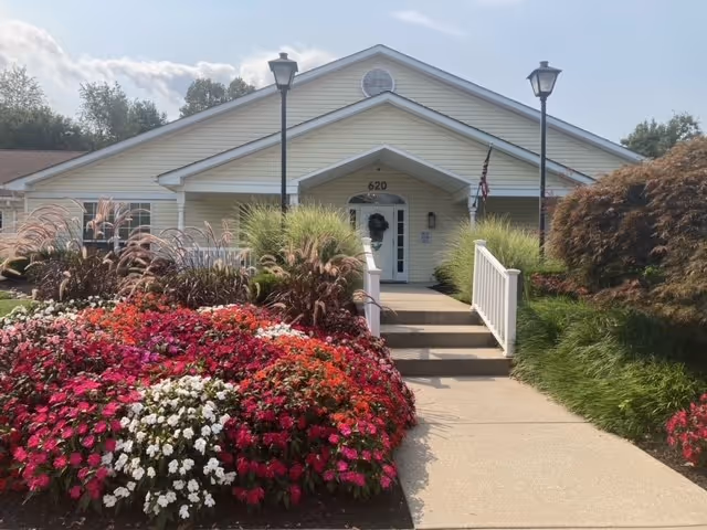 Front exterior view of a single-story building with light yellow siding and a gable roof. The entrance has a small porch with white railings and steps leading up to a white door with the number 620 above it. There are two black lamp posts on either side of the walkway. The foreground features a well-maintained garden with colorful flowers and ornamental grasses.