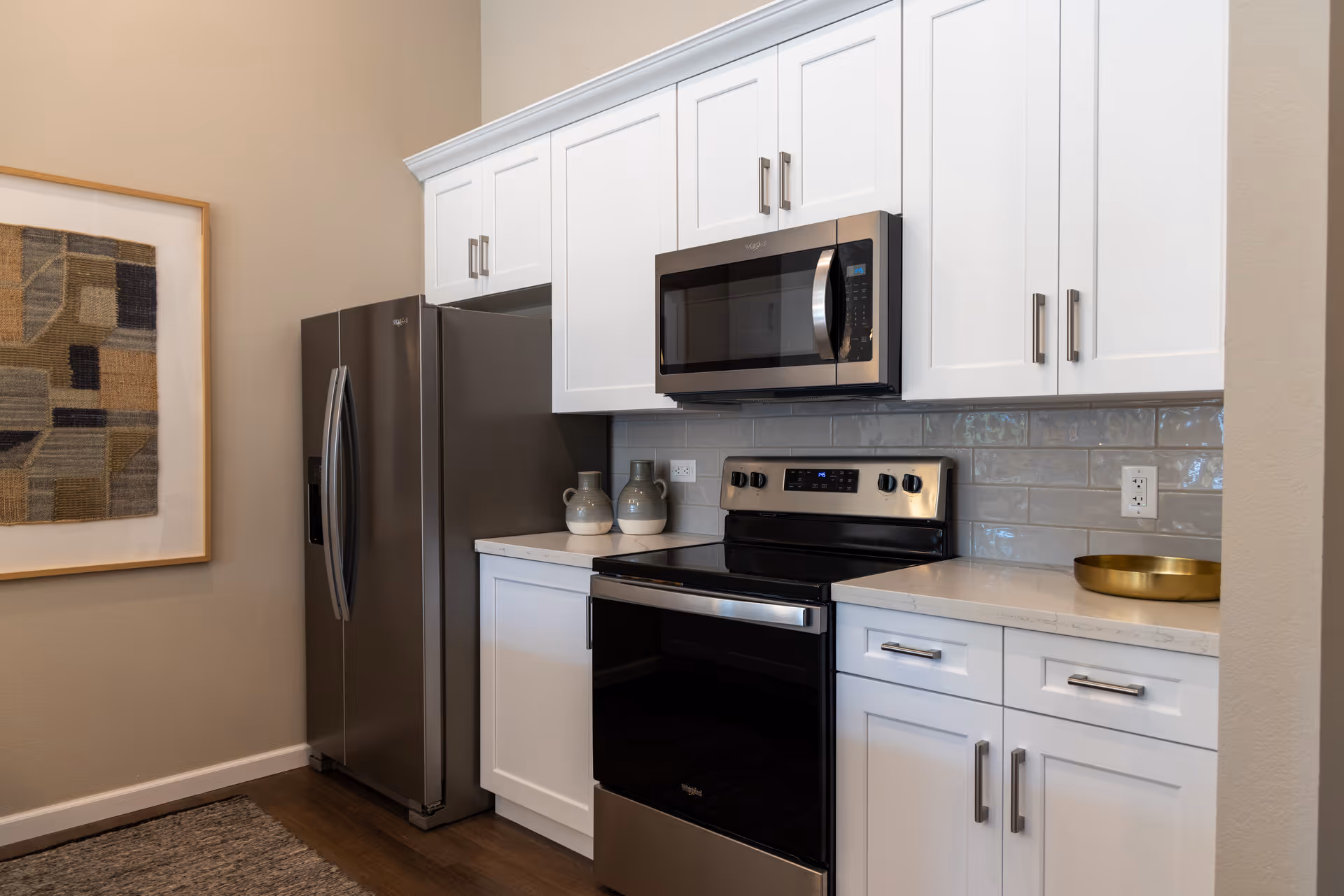Modern kitchen with white cabinets, stainless steel refrigerator, oven, and microwave. There are two decorative ceramic pitchers on the countertop and a brass tray on the right side. A framed abstract textile art piece hangs on the beige wall to the left.