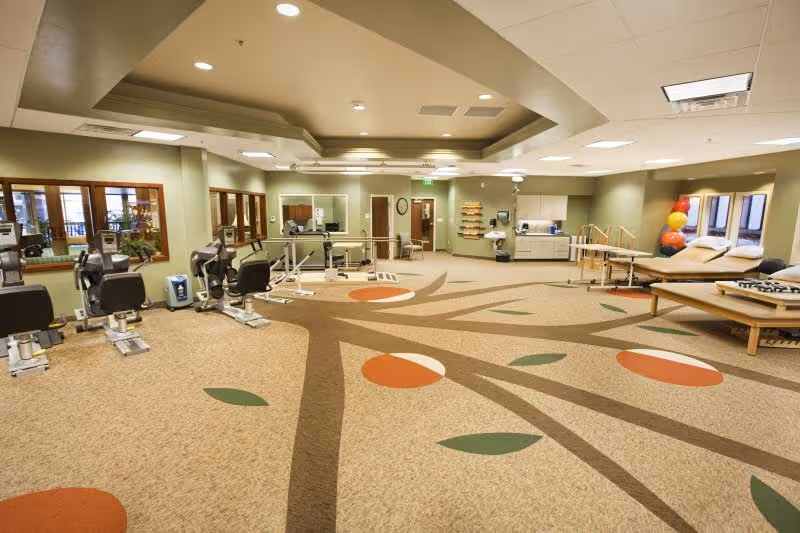 Spacious rehabilitation and fitness room with exercise equipment, therapy tables, and a patterned carpet in a senior living facility.