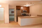 Interior view of a senior living community apartment showing a kitchen area with wooden cabinets, stainless steel refrigerator, microwave, and stove. The kitchen has a countertop with a sink and a breakfast bar. Adjacent to the kitchen is an open doorway leading to another room with carpeted flooring.