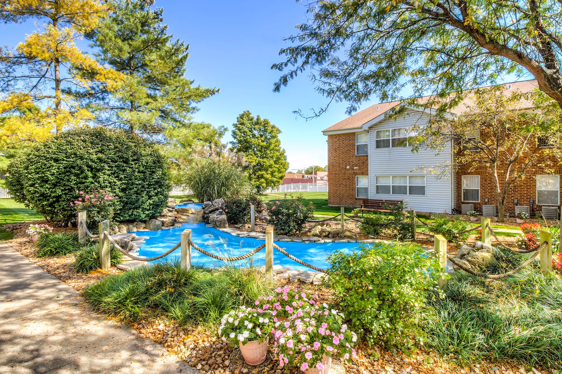 A landscaped outdoor area at a senior living facility featuring a small blue artificial stream bordered by rocks and wooden posts connected with rope. Surrounding the stream are green bushes, flowering plants, and trees. In the background, there is a two-story brick and white siding building with windows and a bench near the entrance. The sky is clear and blue.