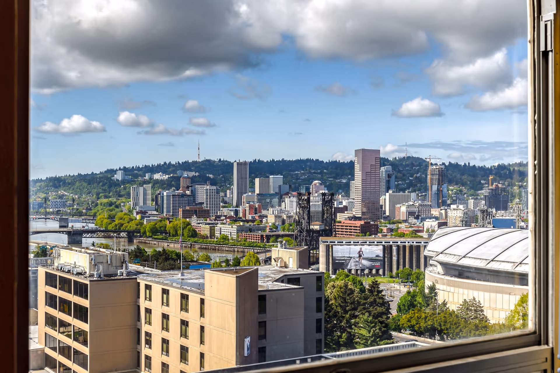 View of a city skyline with various buildings, a river, bridges, and green trees, seen through a window frame under a partly cloudy sky.