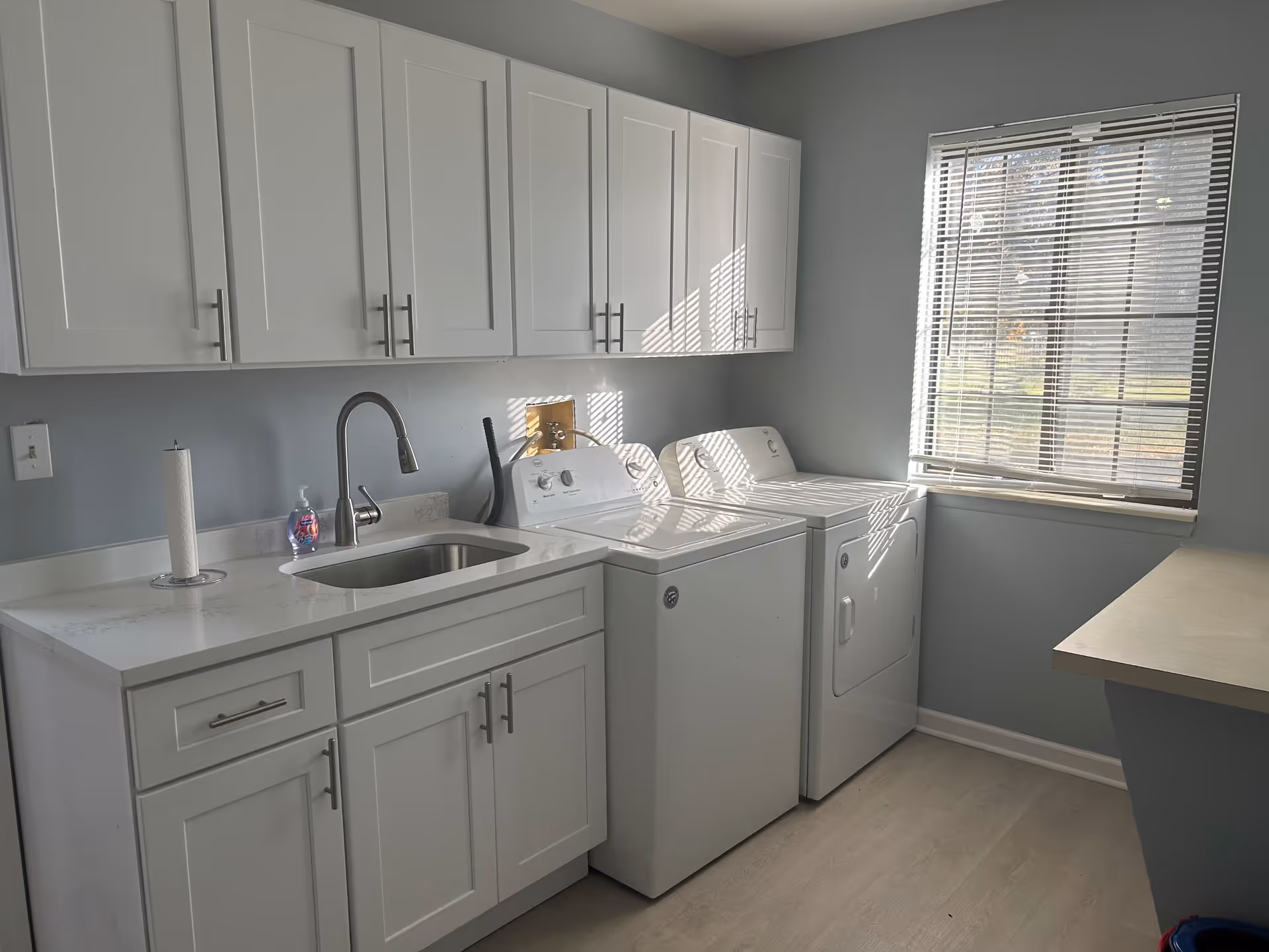 A bright laundry room with white cabinets above and below a countertop. There is a stainless steel sink with a modern faucet, a paper towel holder, and a bottle of hand soap on the counter. Next to the sink are a white washing machine and dryer. A window with blinds lets in natural light, illuminating the room.