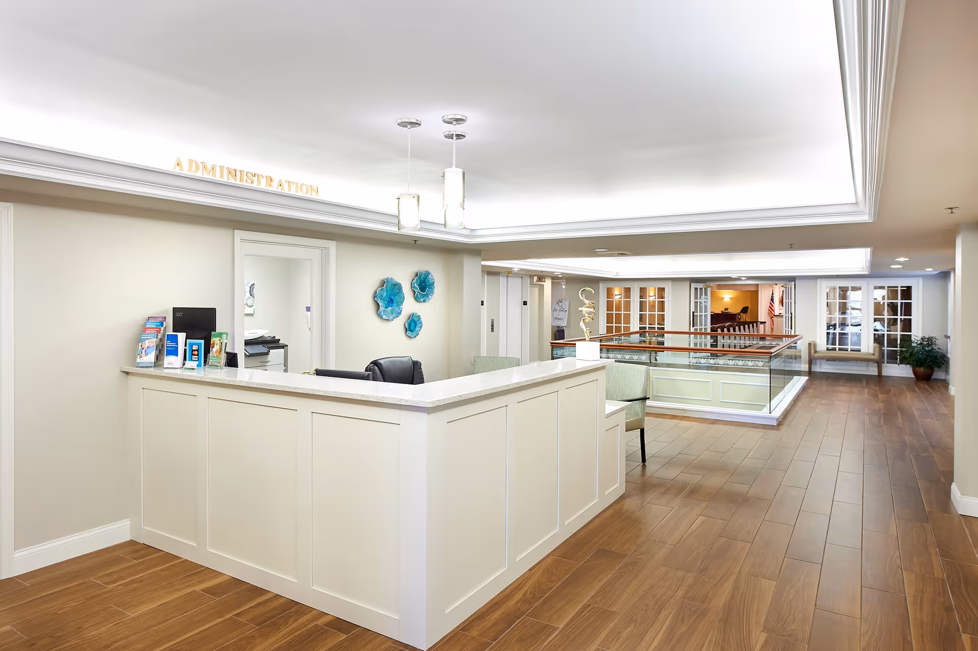 A bright and clean administration area in a senior living facility with a white reception desk, two hanging pendant lights, wooden flooring, and decorative blue wall art. The space extends into a hallway with glass doors and seating areas.