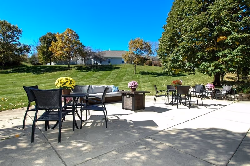 Outdoor patio area with several black metal tables and chairs, decorated with potted flowers. In the background, there is a grassy hill with trees and a building under a clear blue sky.