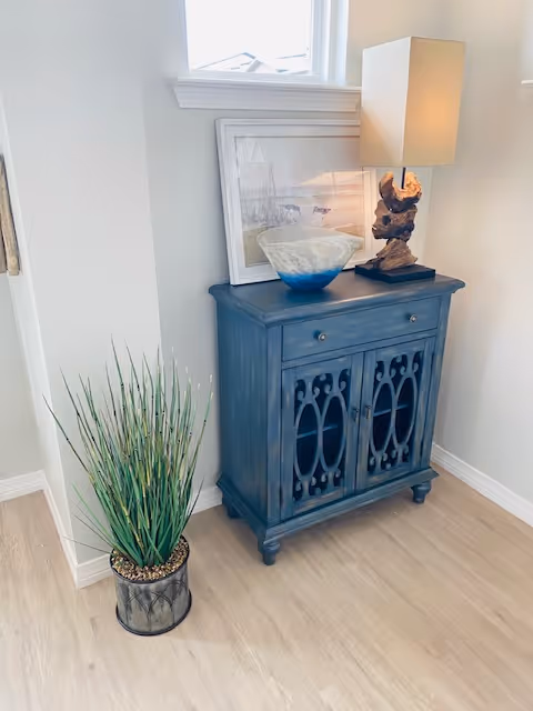 A small blue wooden cabinet with decorative cutouts on the doors, topped with a table lamp with a sculptural base, a framed picture, and a decorative bowl. Next to the cabinet is a potted plant with tall green grass-like leaves. The setting is a corner of a room with light-colored walls and wood flooring.