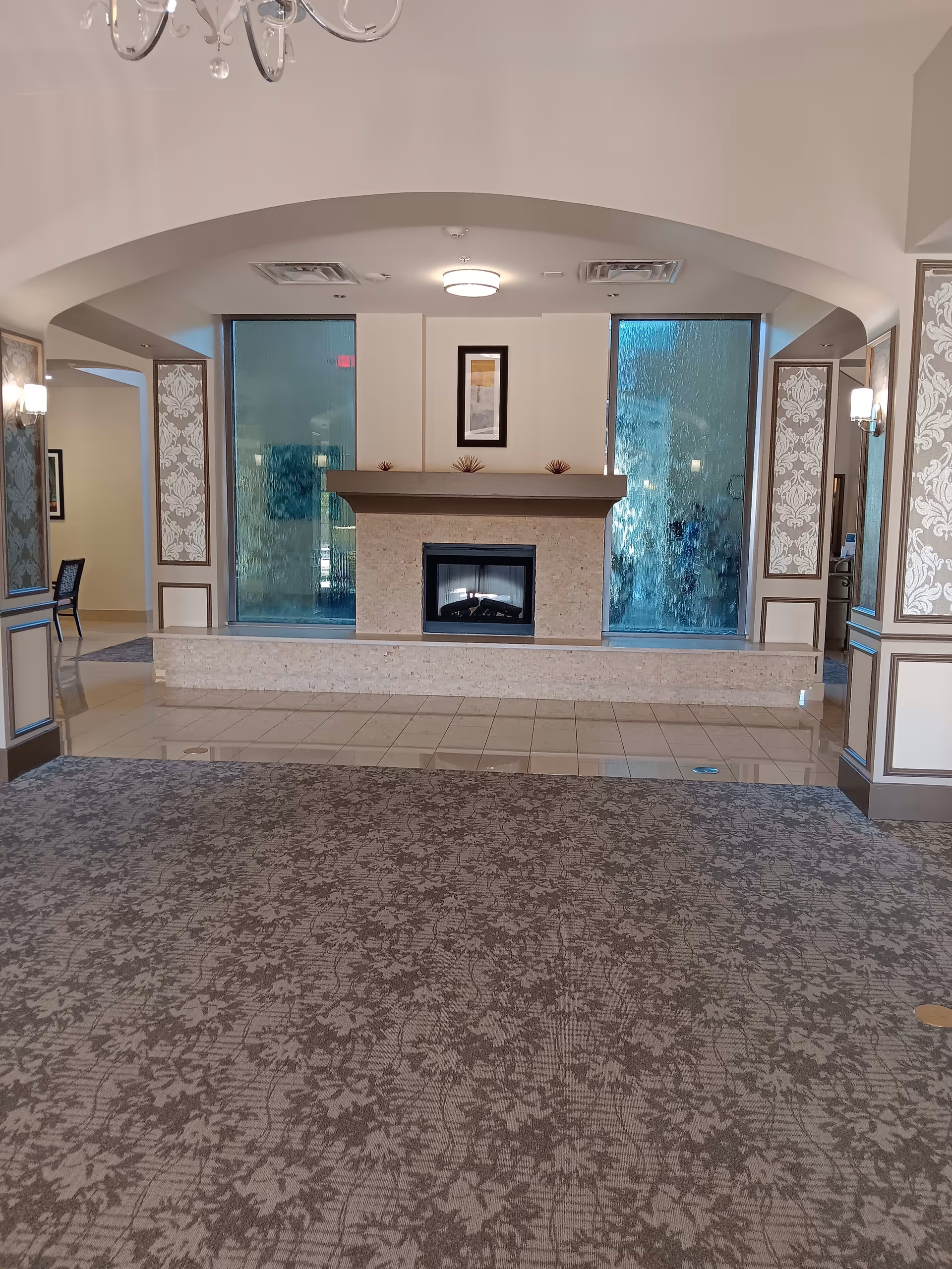 Interior view of a senior living facility lounge area with a decorative fireplace centered between two large textured glass panels. The room features patterned carpet, tiled flooring near the fireplace, wall sconces, and an archway ceiling detail. A framed artwork hangs above the fireplace.