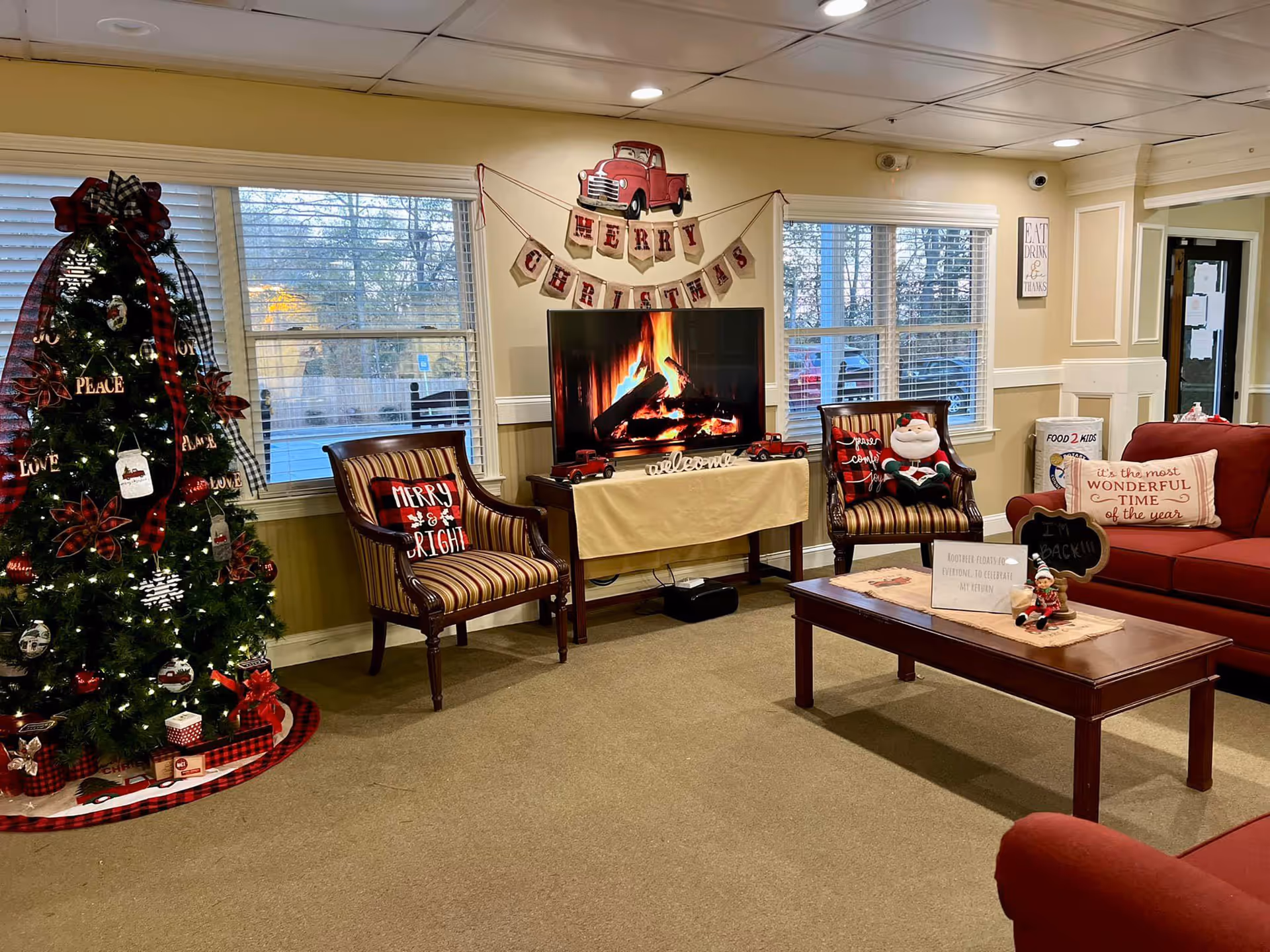 A cozy senior living community lounge decorated for Christmas with a Christmas tree adorned with red and white ornaments and ribbons, two striped armchairs with holiday-themed pillows, a TV displaying a virtual fireplace, and a wooden coffee table with festive decorations. The walls have windows with blinds, holiday banners, and framed signs.