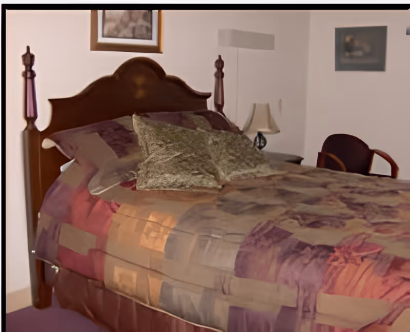 A bedroom with a wooden bed frame featuring a high headboard and two decorative posts. The bed is made with a patterned bedspread in shades of purple, orange, and beige, and has two green textured pillows. There is a small bedside table with a lamp and a wooden chair with armrests in the corner. Two framed pictures hang on the white walls.