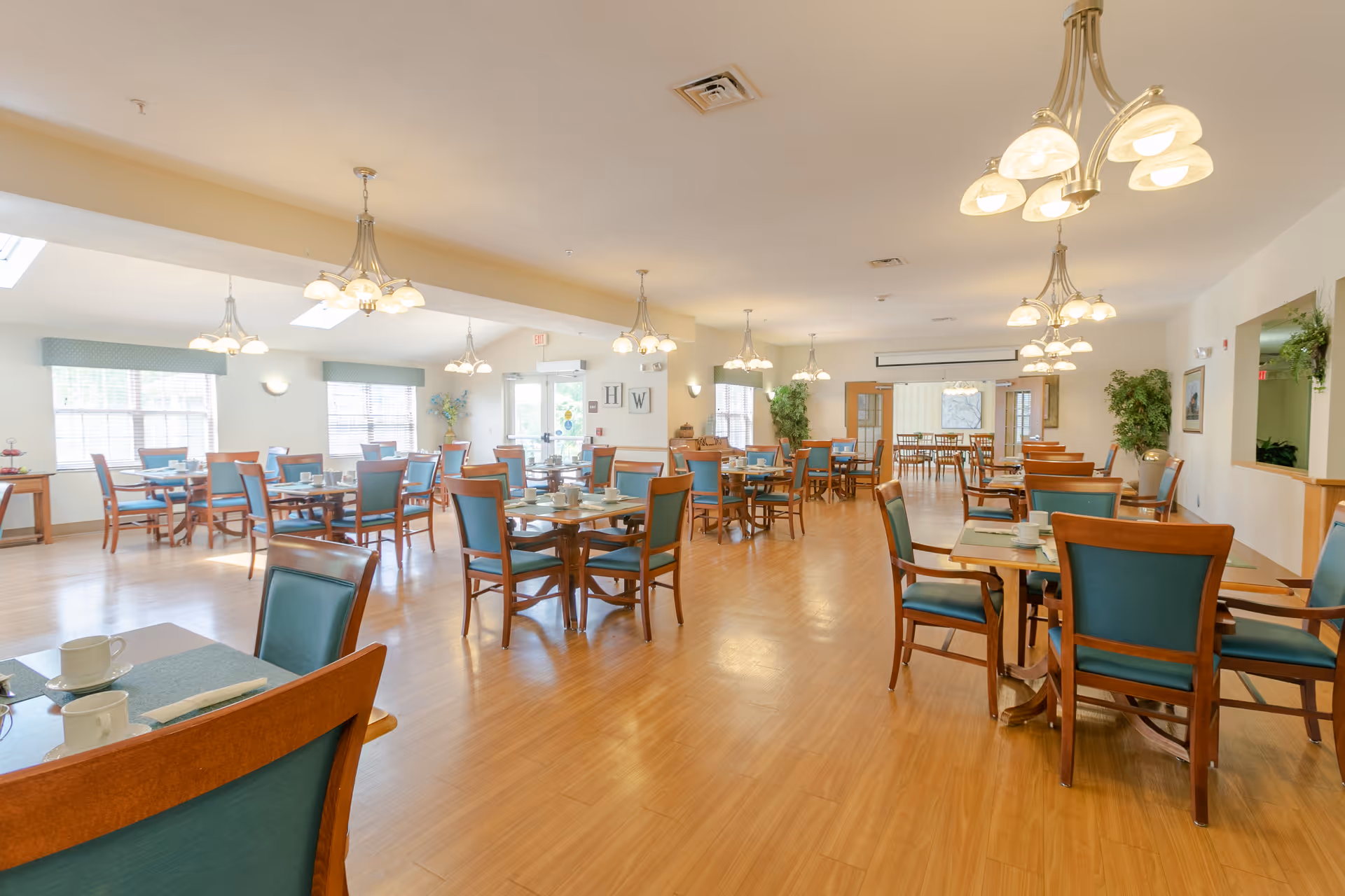 A spacious dining room with multiple wooden tables and chairs with teal cushions. The room is well-lit with natural light from large windows and ceiling lights. The floor is wooden, and there are plants and wall decorations visible in the background.