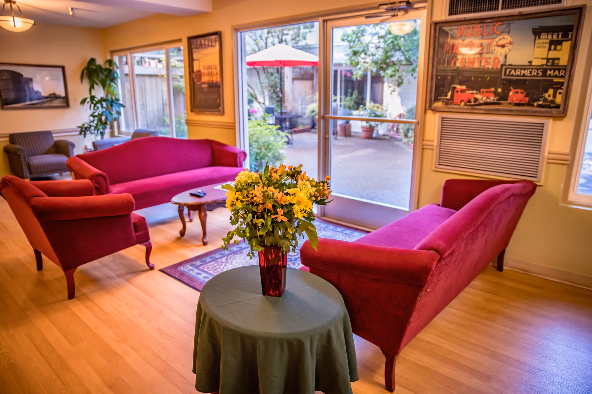 A cozy living room area with red upholstered sofas and armchairs arranged around a wooden coffee table. A small round table with a green tablecloth and a vase of yellow and orange flowers is in the foreground. Large glass doors open to an outdoor patio with plants and a red umbrella. The room has wooden flooring, framed pictures on the walls, and an air conditioning unit below one of the pictures.