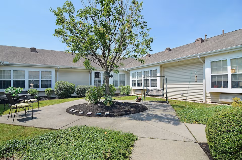 Outdoor courtyard area at Celebration Villa of Lebanon PA featuring a central tree surrounded by a circular mulch bed, concrete walkways, green grass, bushes, and patio chairs. The building exterior has beige siding and multiple windows under a clear blue sky.