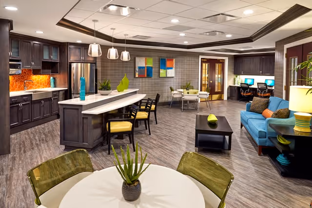 Open communal living area featuring a kitchen island with bar stools, dining tables, a blue sofa, and computer stations.